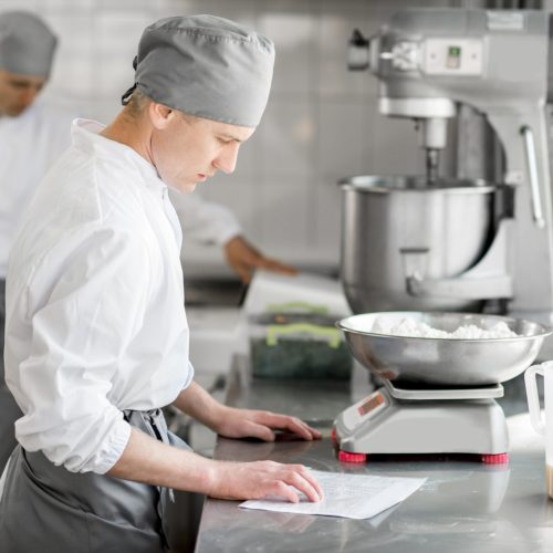 Two handsome male confectioners in uniform weighing ingredients for baking at the manufacturing